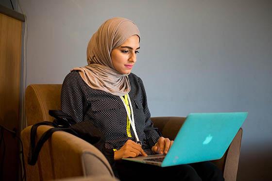 一位年轻的头巾妇女坐在椅子上，膝上放着一台笔记本电脑，正在工作