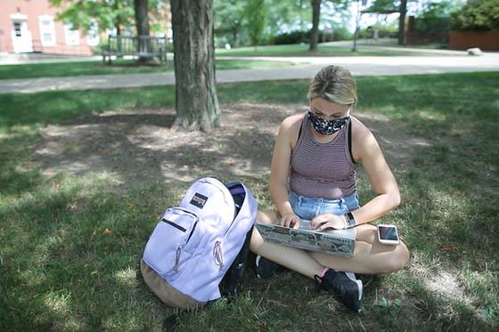 图为一名蒙面学生坐在阴凉校园的地上，用笔记本电脑工作