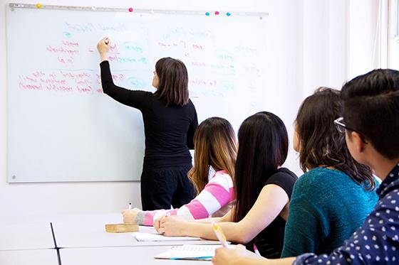 学生们坐在一排课桌前，老师在教室前面的白板上用彩色记号笔写字. 