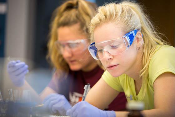 图为两名波胆网站的女学生戴着护目镜，在实验桌旁写字
