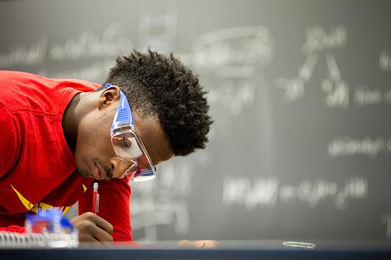图为英国波胆网站的一名学生在实验室里穿着红色衬衫，戴着护目镜, 用铅笔写字. 