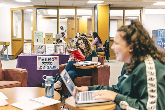 Photo of Chatham University students gathering in Jennie Mellon King Library, sitting in chairs 和 tables while working on laptops 和 reading books. 