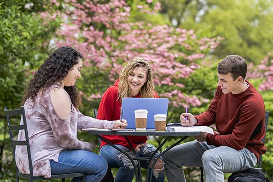 图为三名波胆网站的学生坐在户外露台的桌子旁，一起用笔记本电脑工作，面带微笑. 