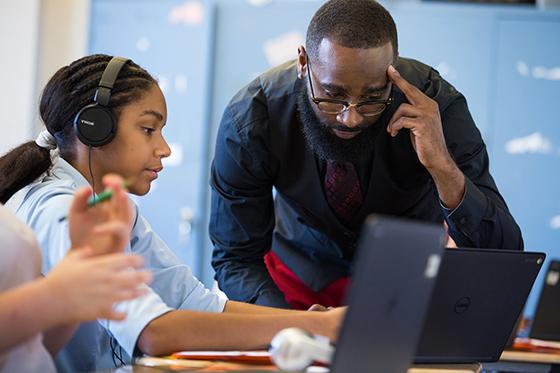 图为在计算机实验室里，一位教授戴着黑色耳机，俯身看着正在用笔记本电脑工作的学生. 